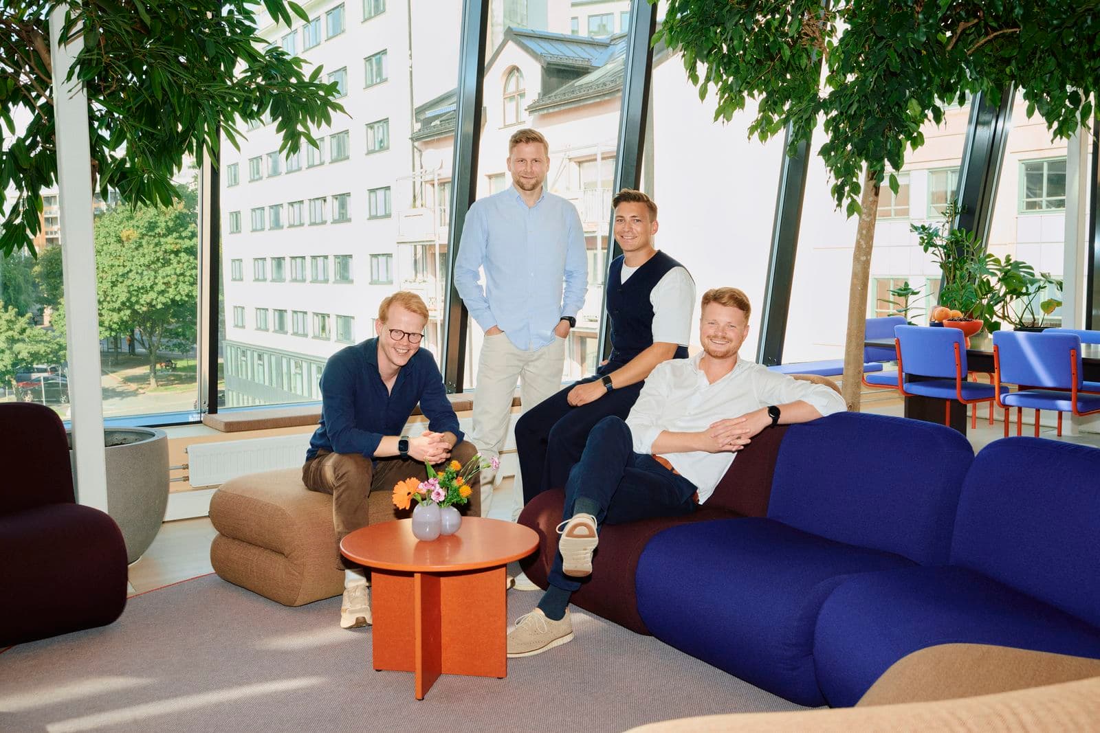 Four men smiling in a modern lounge with large windows, surrounded by plants and colorful furniture.